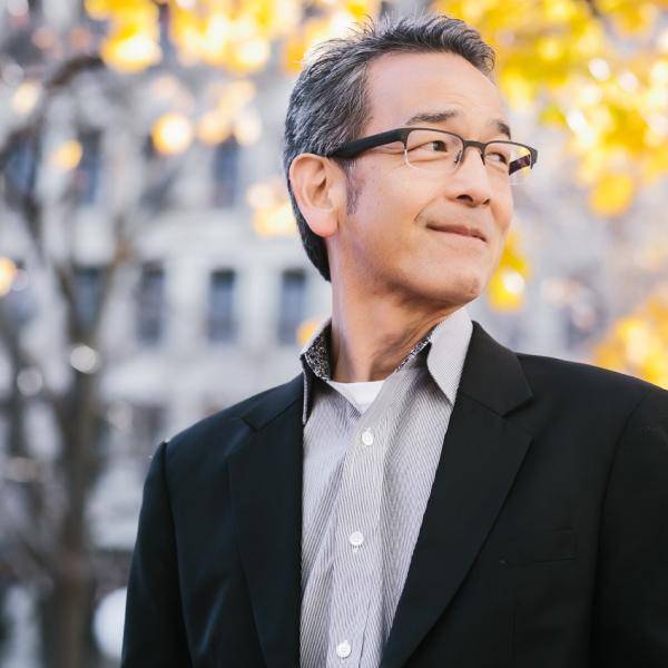 A photograph of David Nagahiro in front of a blurred background consisting of a building and orange foliage. He is smiling and looking to his right. He is wearing glasses, a black suit jacket, and a grey dress shirt.