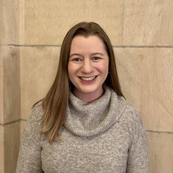 Maria is smiling at the camera, she is wearing a white sweater, she has long hair.